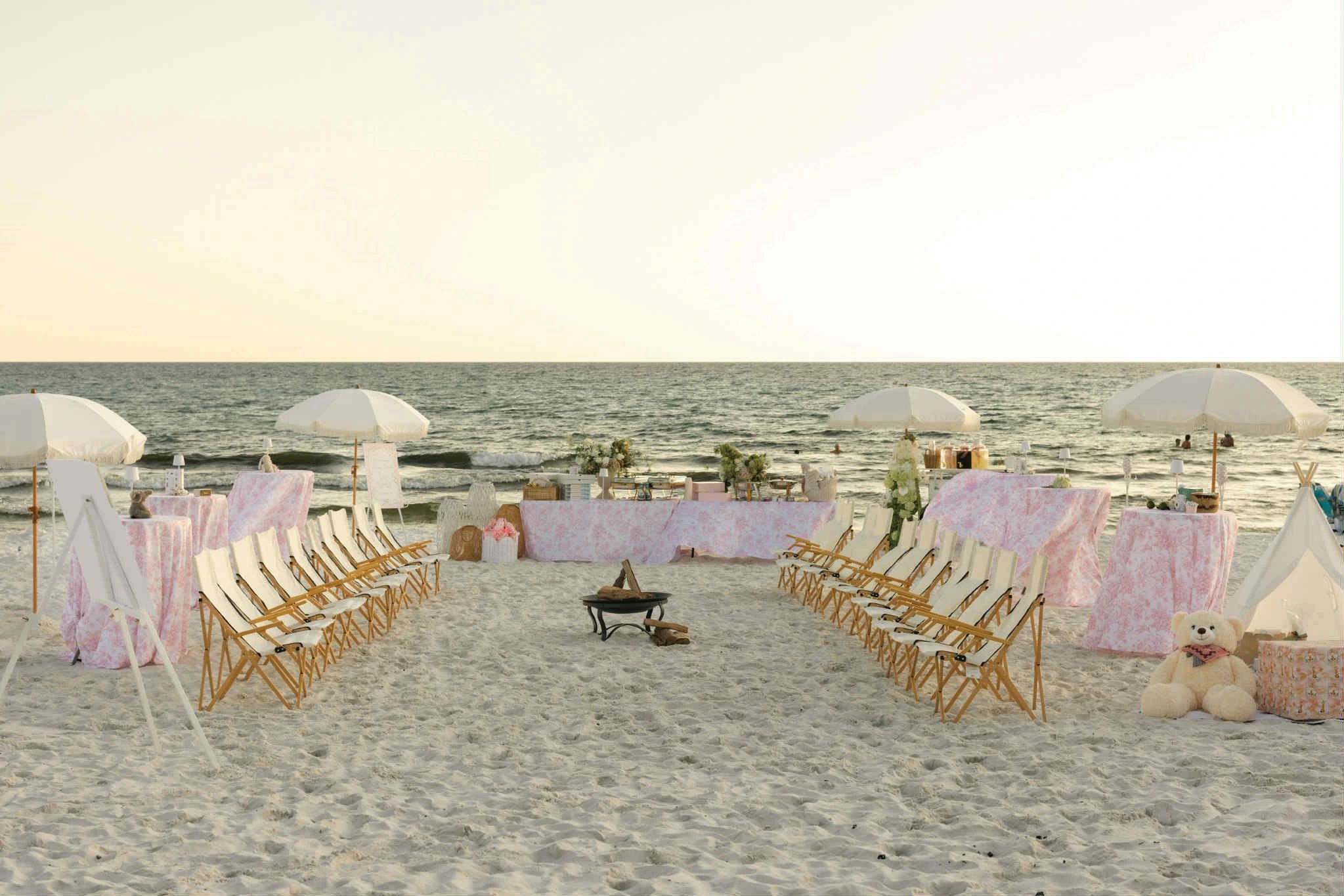 Beach event setup with chairs, umbrellas and a fire pit on the sand